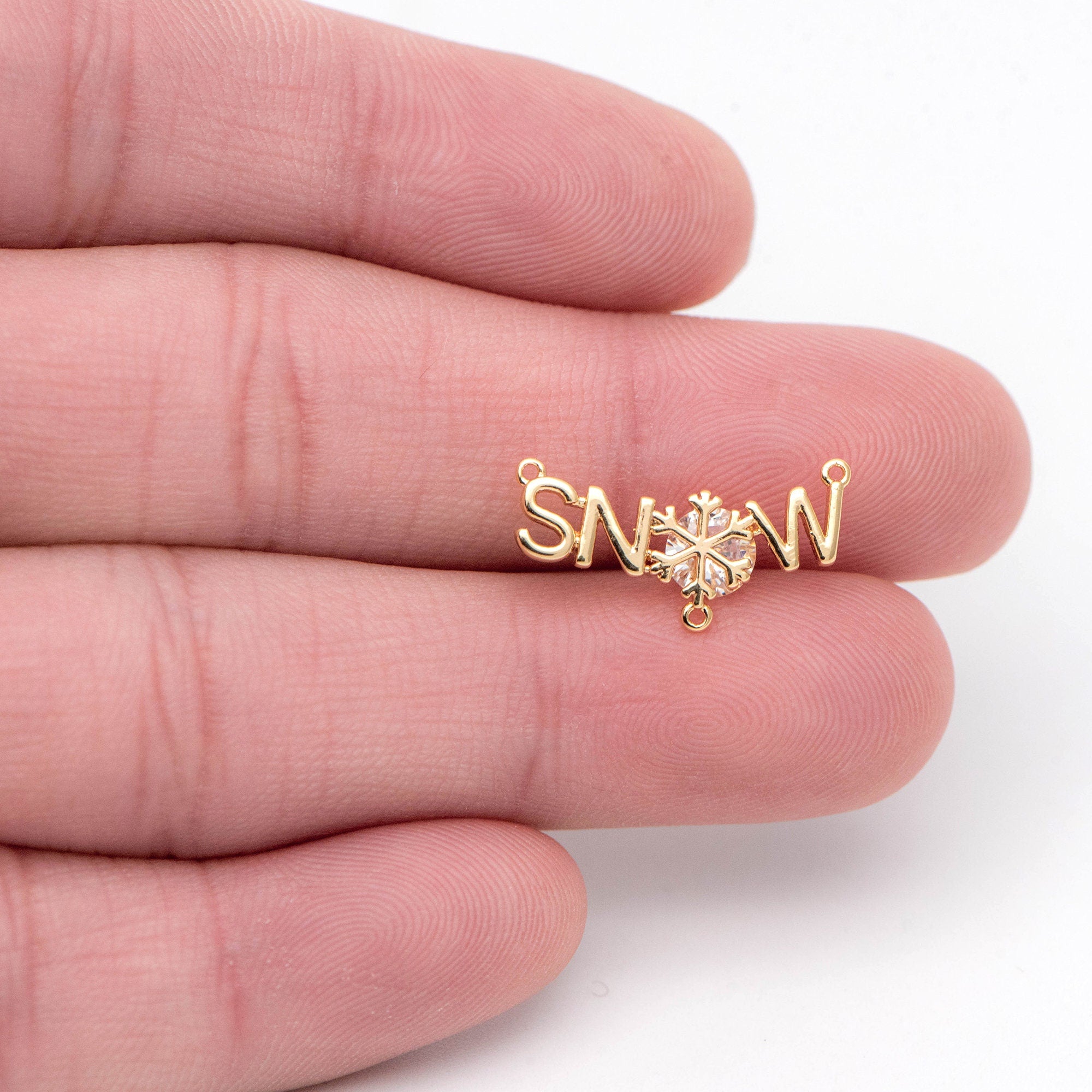 May include: A gold charm with the word 'SNOW' spelled out in cursive letters. A snowflake with a clear rhinestone center is in the center of the charm. The charm has two jump rings for attaching to a necklace or bracelet.